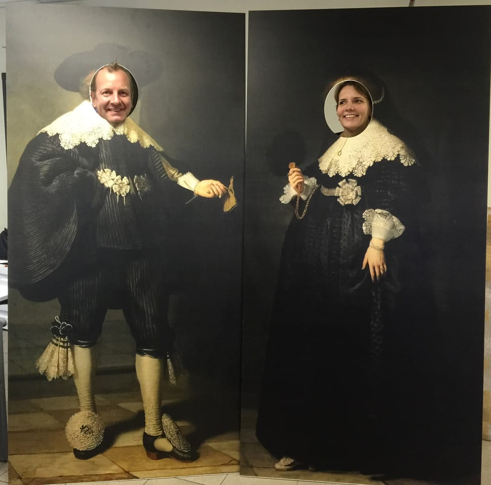 Twee fotoborden met een klassiek schilderij als motief en uitgesneden gezichtsgaten, met twee mensen erachter