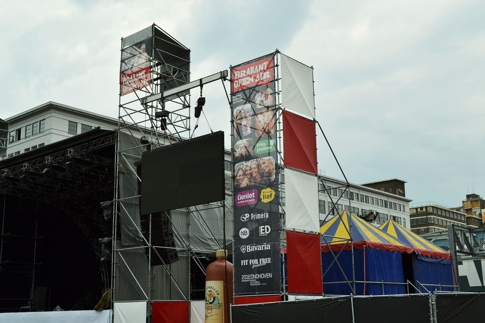 Meshdoeken met festivalprogramma aan een steigertoren naast het hoofdpodium van een openluchtfestival